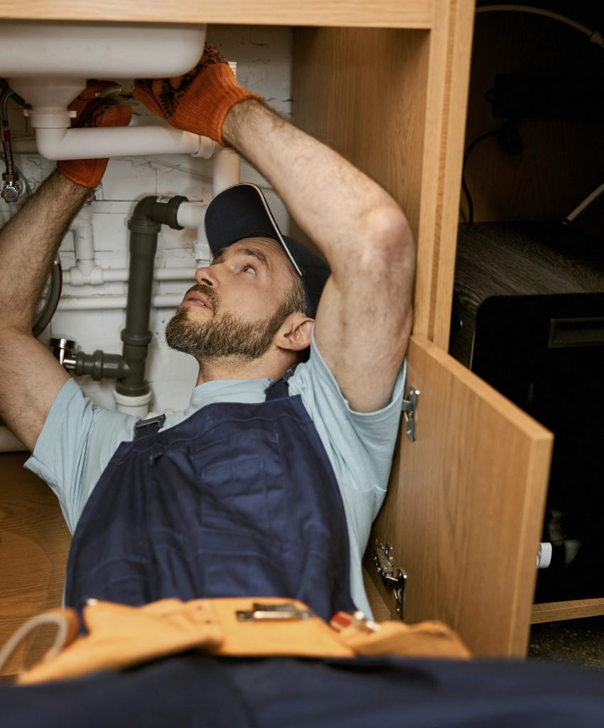 Bearded young man wearing work gloves and cap while fixing kitchen sink
